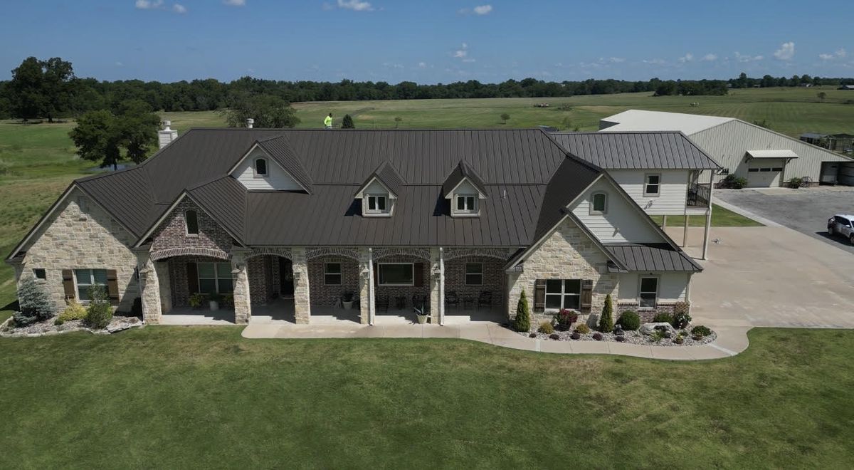 Aerial view of a stone ranch home with a dark metal roof and a large lawn